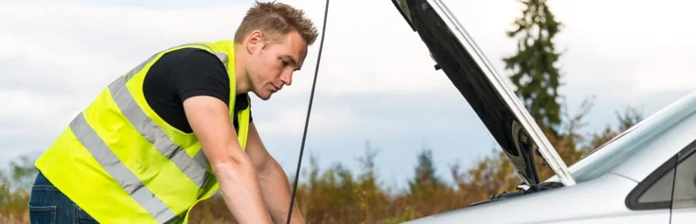 Homem jovem, com um colete refletor vestido, observa o motor do seu carro avariado.