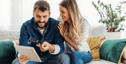 Casal jovem sentado no sofá verifica tablet Casal jovem, sentado num sofá, a olhar para um tablet e a sorrir