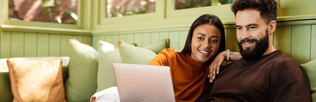 Casal sorridente faz um seguro de vida online, sentado no sofá com portátil nos joelhos.