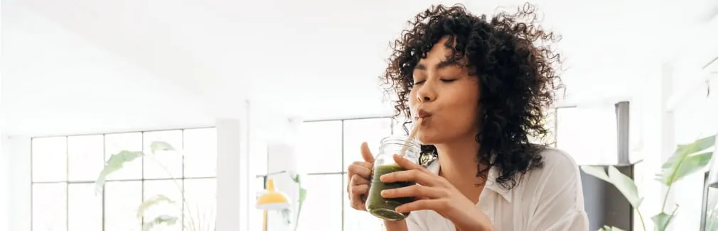 Mulher jovem, apoiada numa bancada de madeira, bebe um batido para desintoxicar o corpo.
