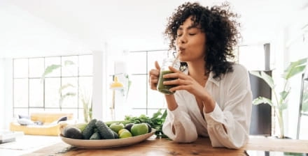 Mulher jovem, apoiada numa bancada de madeira, bebe um batido para desintoxicar o corpo.